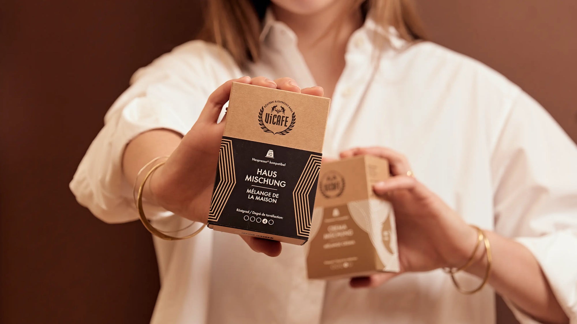 Woman holding two boxes of ViCACE Specialty coffee capsules against a brown background